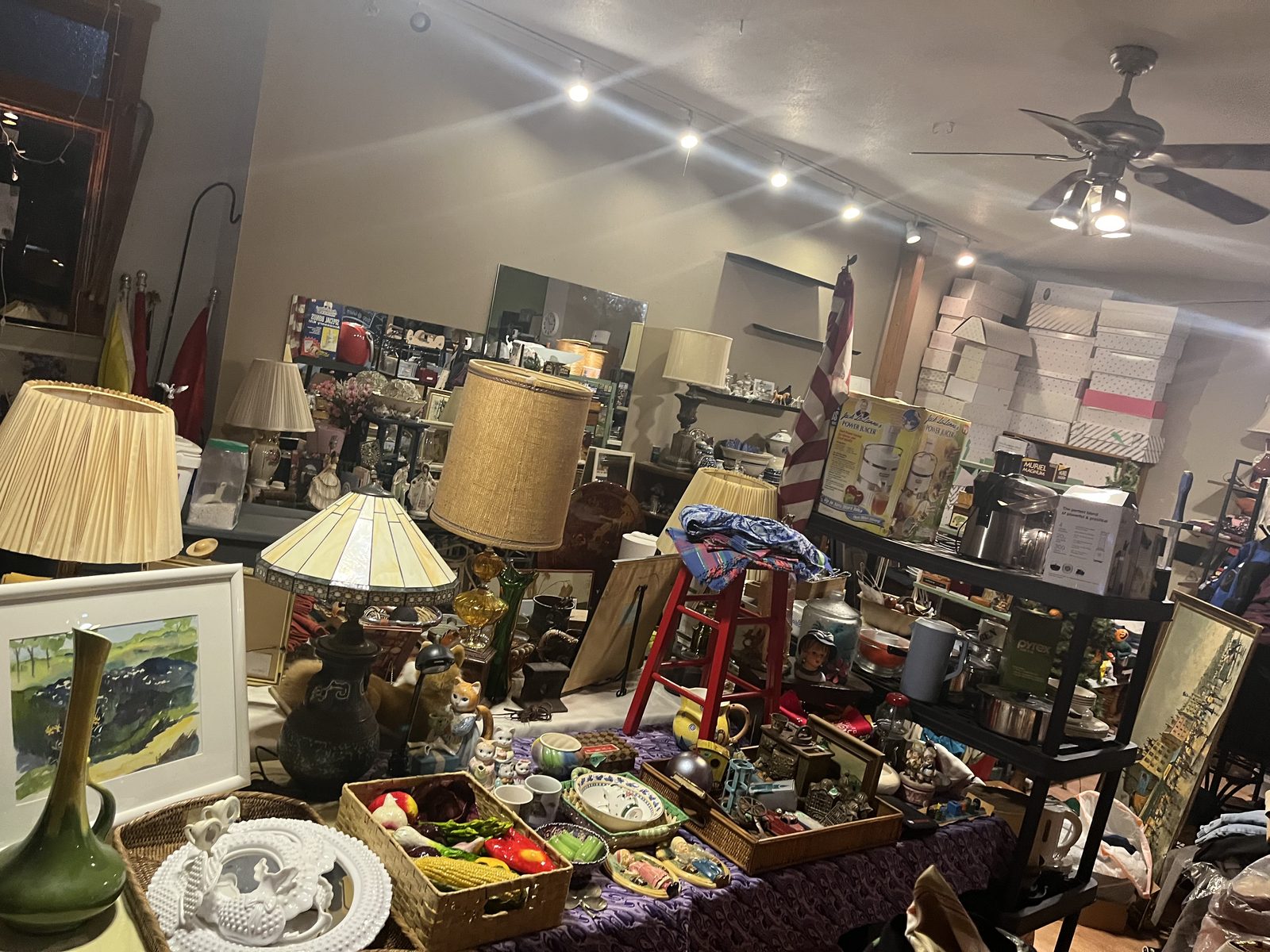 Sale floor at Simple Treasures during setup: table lamps, a framed watercolor, baskets of ceramic fruit and vegetables, and shelves of collectibles