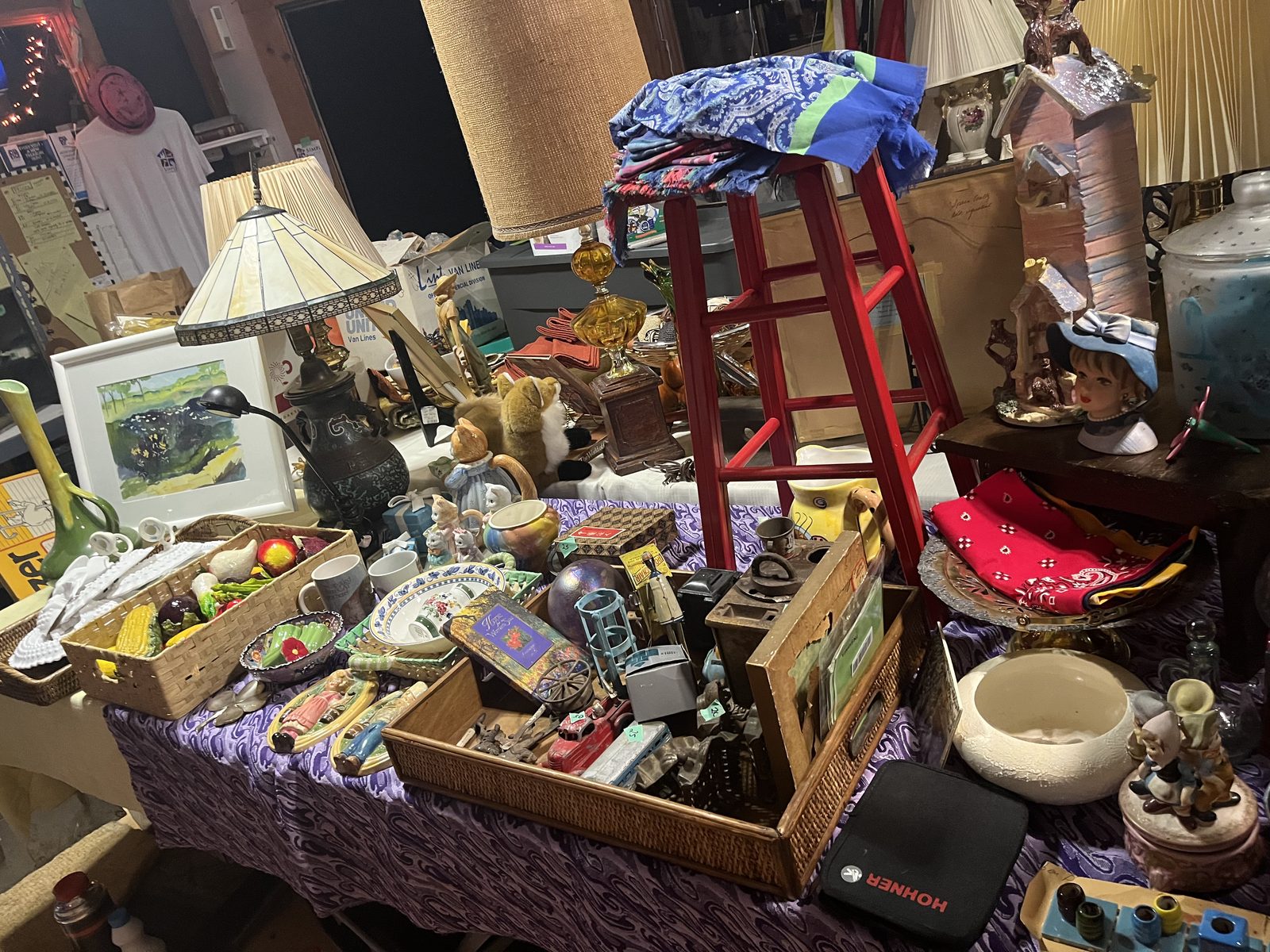 Sale table with baskets of ceramic vegetables, a stained glass lamp, a red stool, a lady head vase and a framed watercolor